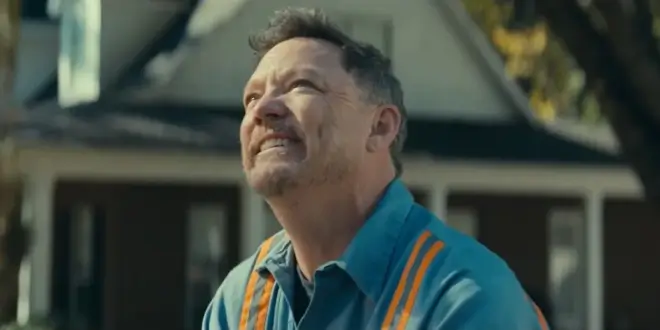 Matthew Lillard looks up smiling while wearing a blue work shirt with orange stripes in The Life of Chuck.