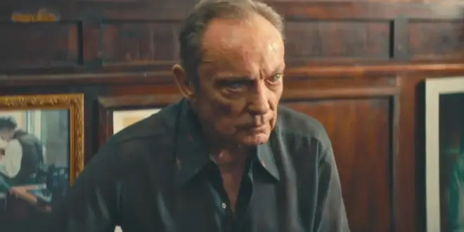 Udo Kier sits with cryptic worry in a wood paneled room in The Secret Agent.