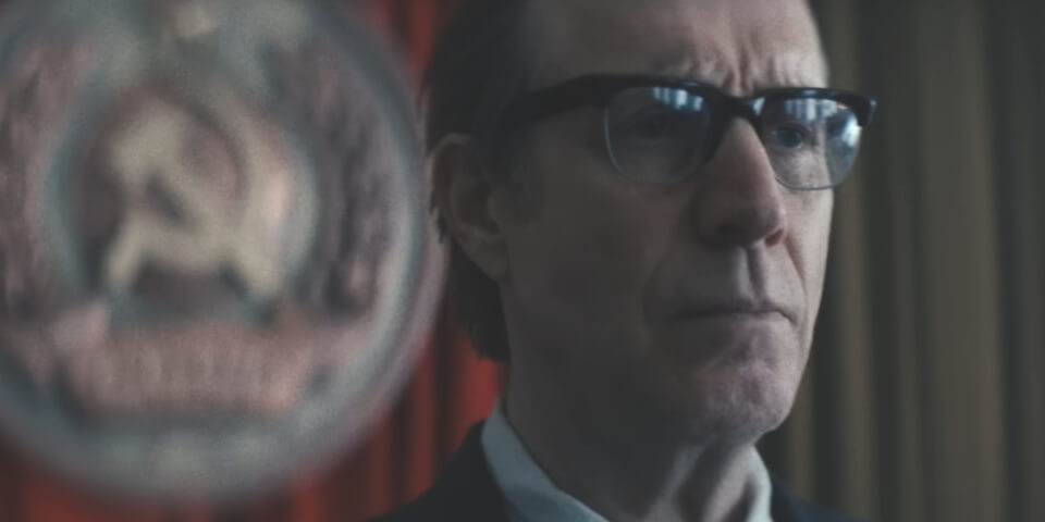 A solemn looking Rhys Ifans stands in a meeting hall in Star City.