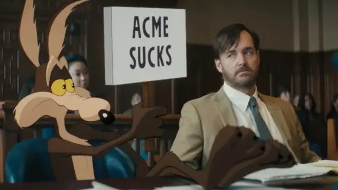 Wile E. Coyote holds up an anti ACME sign as Will Forte looks on with concern in Coyote vs. Acme.