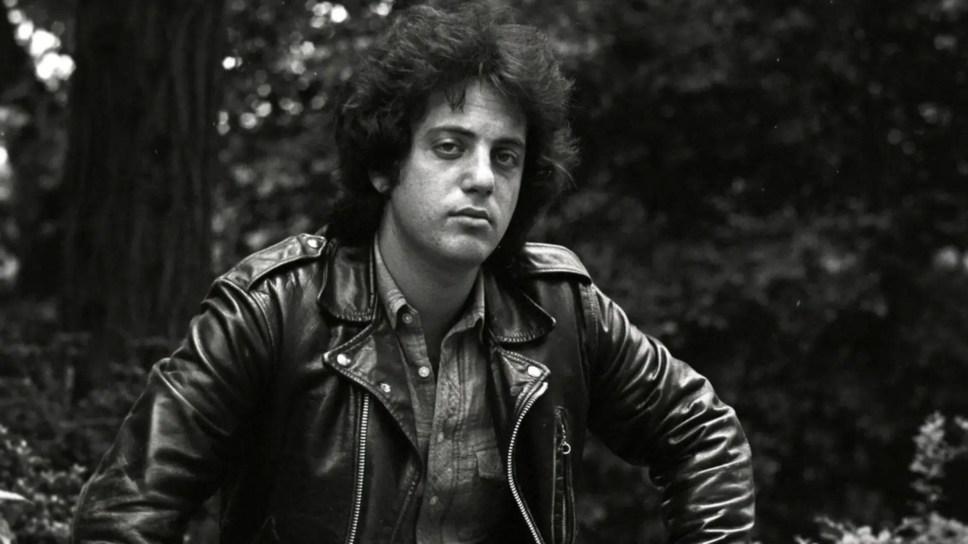A young Billy Joel sits on a park bench in a leather jacket, in a photo from Billy Joel: And So It Goes' press kit.