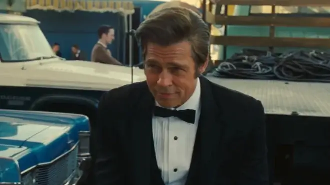 A tuxedoed Brad Pitt sits against a car with a mischievous smile in Once Upon a Time in Hollywood.