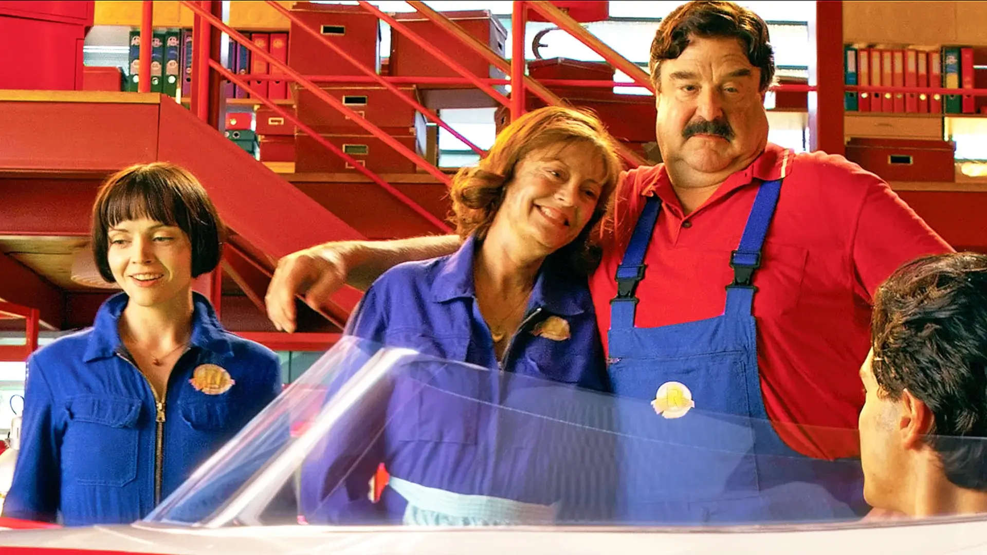Christina Ricci, Susan Sarandon, and John Goodman stand smiling in the garage next to the Mach 6 in Speed Racer.