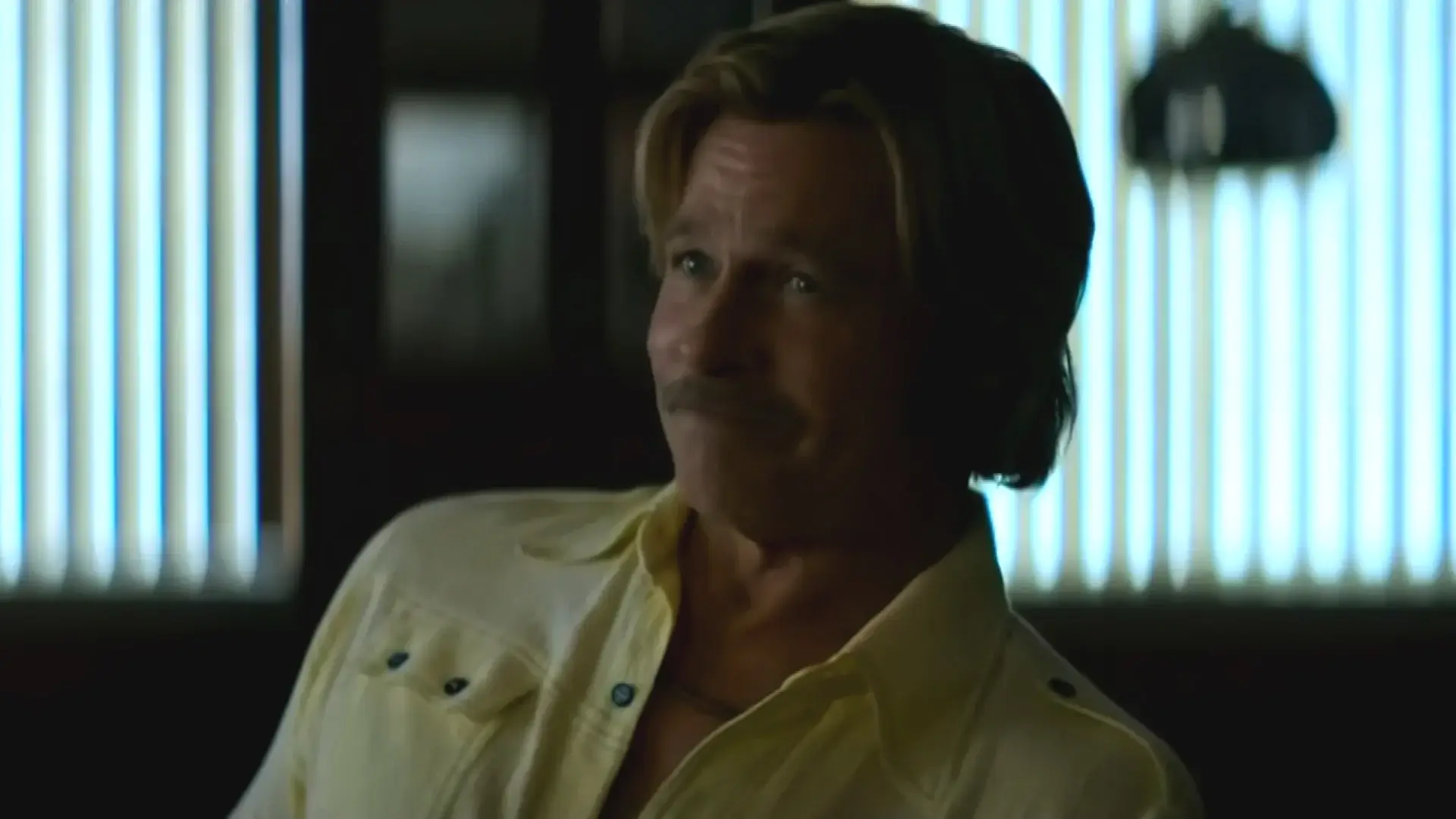 Brad Pitt sits with a cocky smile in a dimly lit office in The Adventures of Cliff Booth.