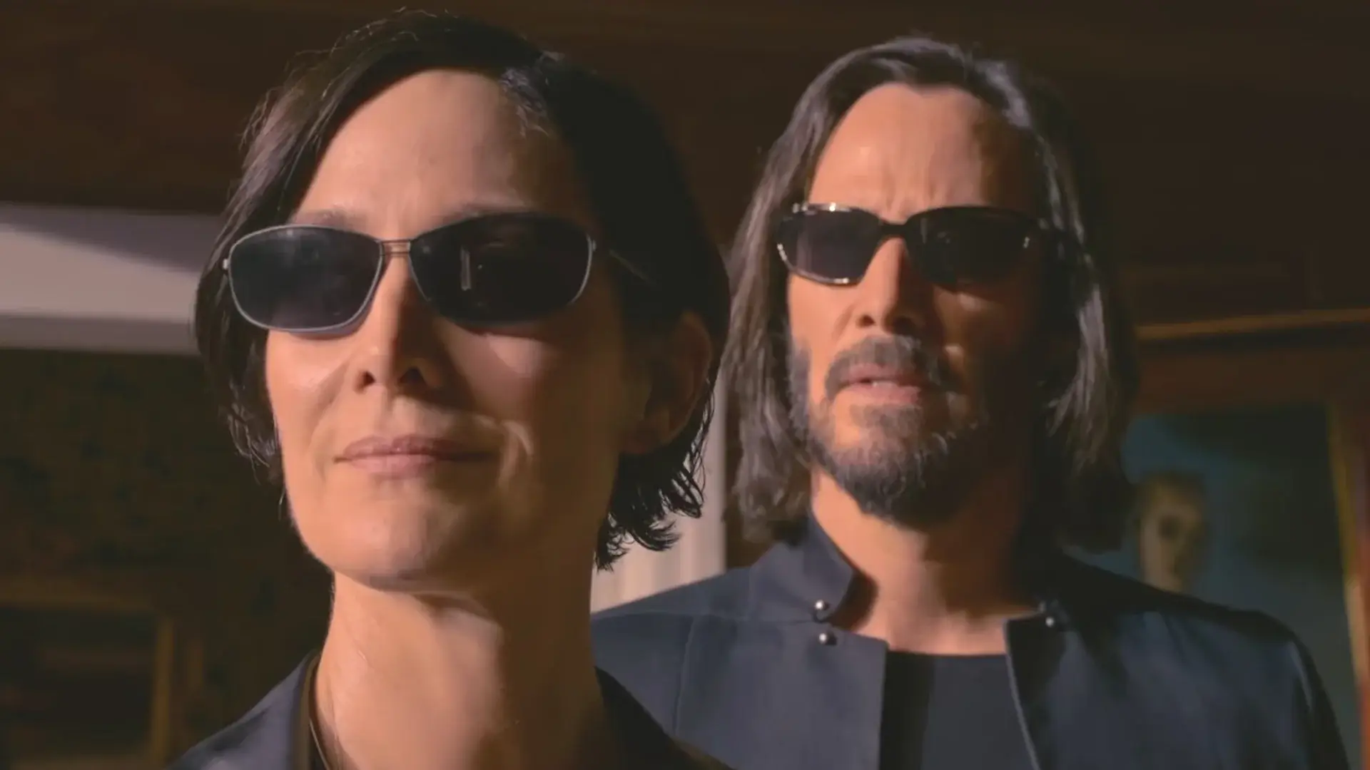 Carrie Anne Moss and Keanu Reeves stand smiling while wearing sunglasses in The Matrix Resurrections.