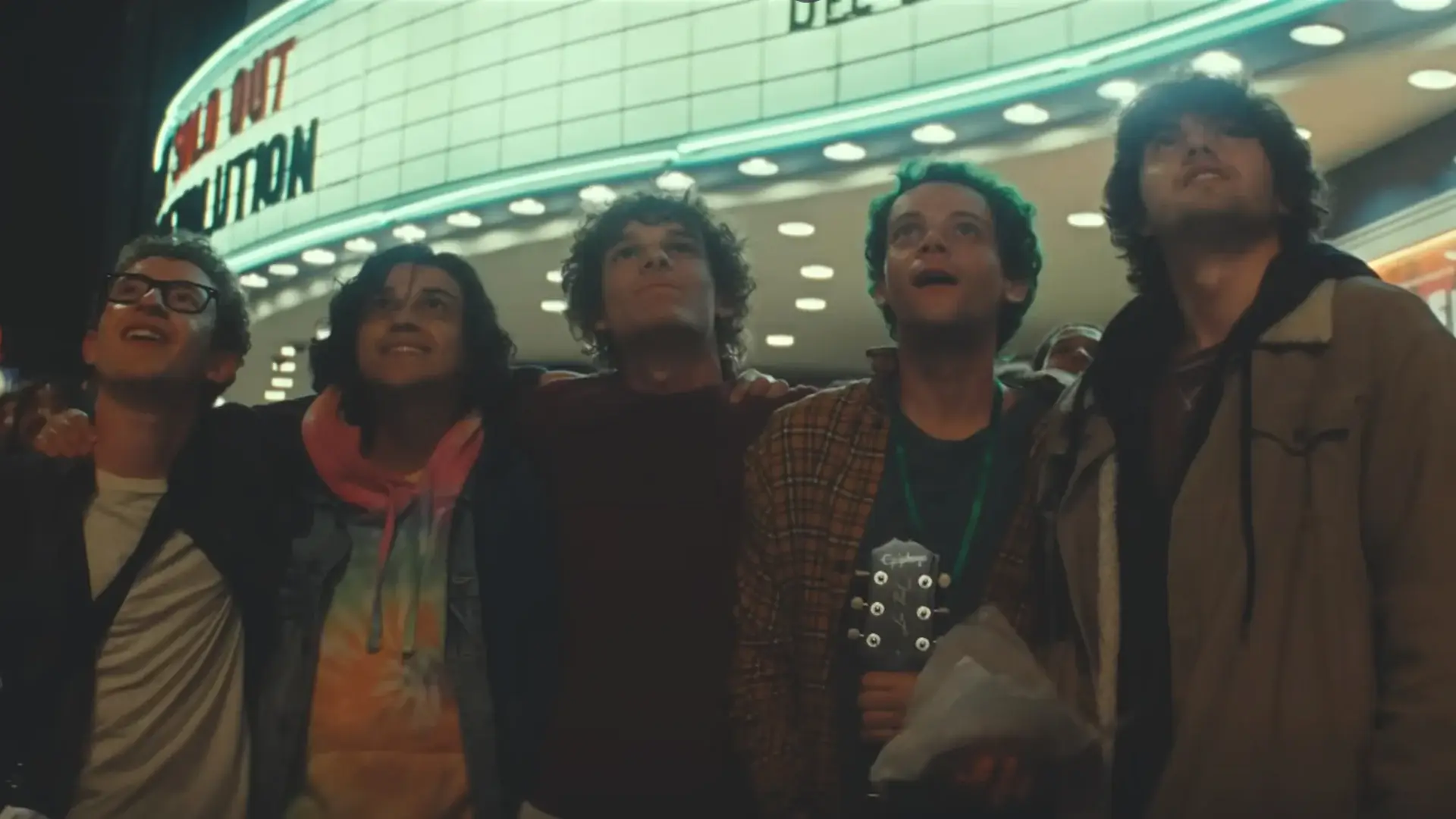 The cast of Nimrods look at the night sky with smiles, while standing in front of a theater marquee