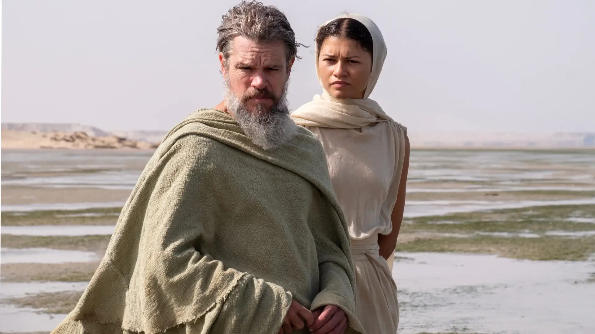 A bearded Matt Damon stands in a desolate location with Zendaya watching from behind in The Odyssey.