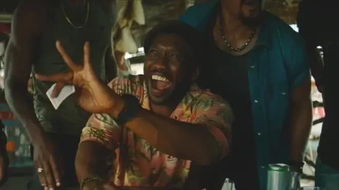 Mahershala Ali smiles while holding up a winning hand of cards in Jurassic World Rebirth.