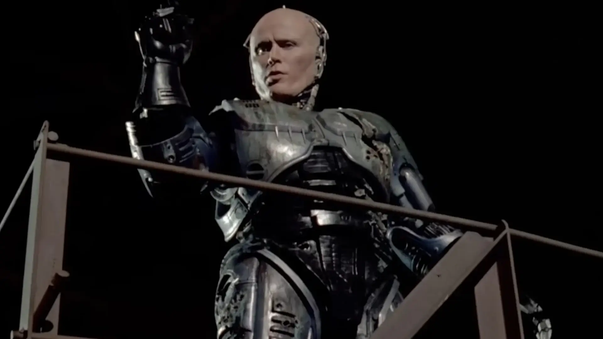 An unmasked Peter Weller stands on a catwalk aiming his gun in the air in Robocop.