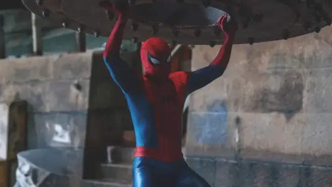 Tom Holland stands in his Spider-Man costume while holding a heavy object over his head in Spider-Man: Brand New Day.