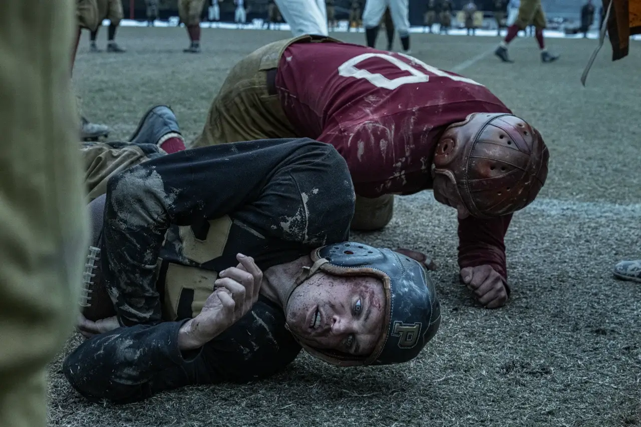 "The Mosquito Bowl. Two vintage football players wearing old-style helmets and uniforms lie on a muddy field after a tackle, both looking exhausted; blurred players and spectators are seen in the background during a gritty, historical game."