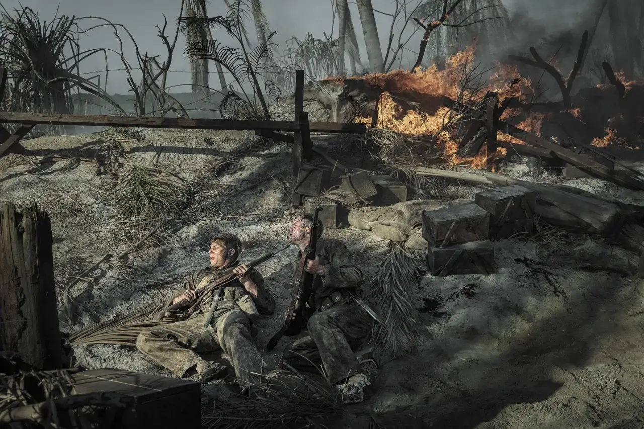 "The Mosquito Bowl. Two soldiers in dirty uniforms lie on a smoky, war-torn battlefield holding rifles, surrounded by debris and crates, as fire rages behind them and palm trees stand amid destruction."