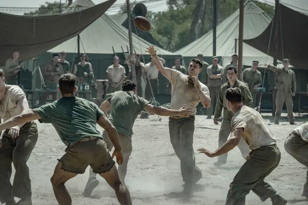 "The Mosquito Bowl Soldiers in vintage uniforms play football on a dusty field surrounded by military tents, with other soldiers watching, suggesting a break in a wartime or military camp setting."