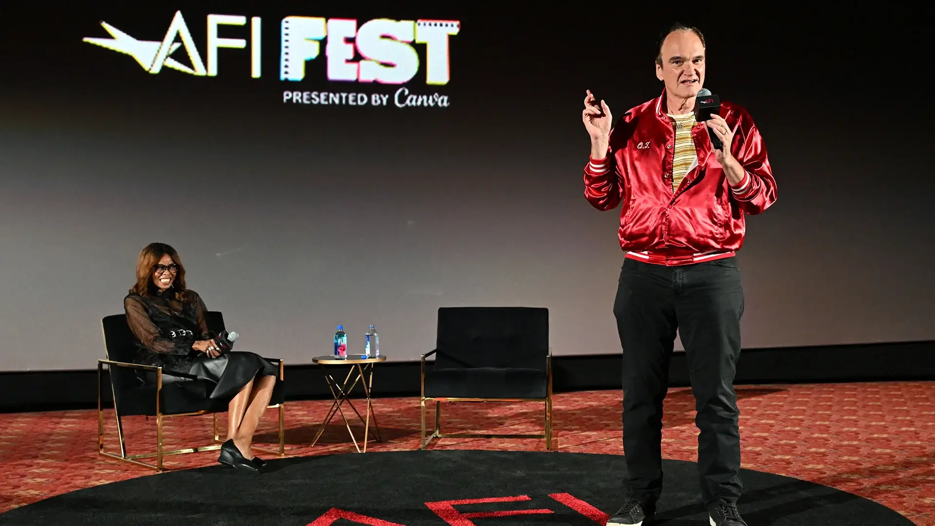 Man in red jacket talks on stage with woman behind him.